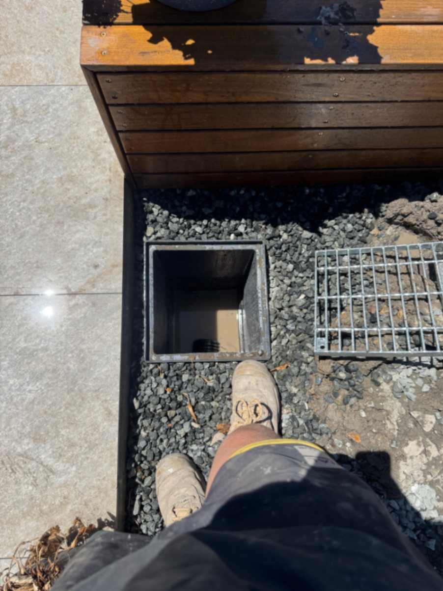 Drainage pit with grate beside wooden deck.